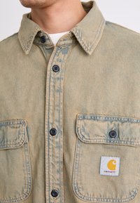 Jasno beżowa koszula dżinsowa Carhartt z guzikiem z przodu, spiczastym kołnierzykiem i dwiema kieszeniami na piersi z metką logo Carhartt.
