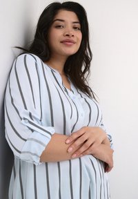Blouse rayée bleu clair avec un col, boutons à l'avant et manches trois-quarts. Rayures verticales grises et texture de tissu douce.