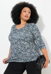 Langärmliges Oberteil in einem schwarz-weiß gesprenkelten Muster mit V-Ausschnitt. Der Stoff wirkt weich; getragen mit schwarzen Hosen und einer schwarzen Clutch.