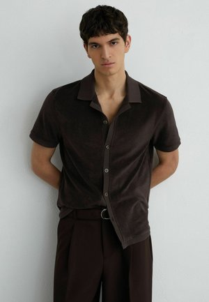 Junger Mann mit kurzen dunklen Haaren, der ein dunkelbraunes kurzärmeliges Hemd und passende dunkle Hose vor einer schlichten hellen Wand trägt.