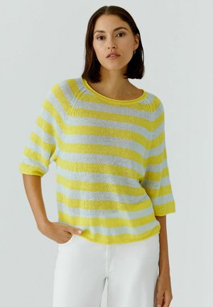 Femme portant un pull loose rayé jaune et bleu clair à manches trois-quarts et un pantalon blanc, debout devant un fond uni.