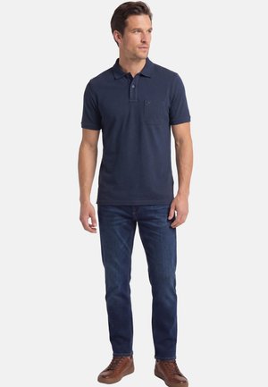 Man draagt een marineblauw poloshirt met korte mouwen, donkerblauwe spijkerbroek en bruine leren sneakers, staand en zijwaarts kijkend.