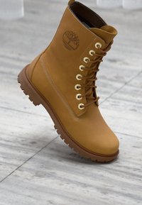 Botas de cuero color marrón, con cordones y puntera redonda, superficie texturizada y suela de goma, que presentan ojales dorados y un logo grabado en el lateral.
