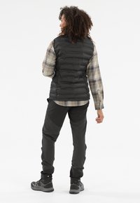 Schwarze Steppweste über einem karierten Langarmhemd, kombiniert mit dunklen Hosen und grauen Wanderschuhen. Das Outfit zeichnet sich durch ein geschichtetes Design aus.