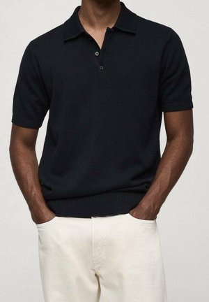 Mand iført en sort strikket polo med korte ærmer og offwhite bukser med hænderne i lommerne, vist fra hals til hofter.