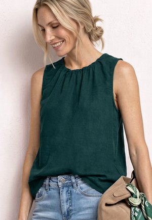 Blonde Frau lächelnd, trägt ärmelloses dunkelgrünes Oberteil und hellblaue Jeans, hält eine hellbraune Wildledertasche mit grünem gemustertem Schal.