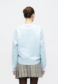 Γυναίκα με μαλλιά σε κότσο, που φοράει ανοιχτό μπλε φούτερ, γκρι καρό πλισέ φούστα και μαύρες κολάν, όρθια με την πλάτη στραμμένη.