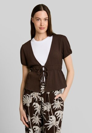 Vrouw met stijl donker haar die een gestructureerde bruine korte mouw top draagt over een wit overhemd en bruine broek met witte palmboomprints.
