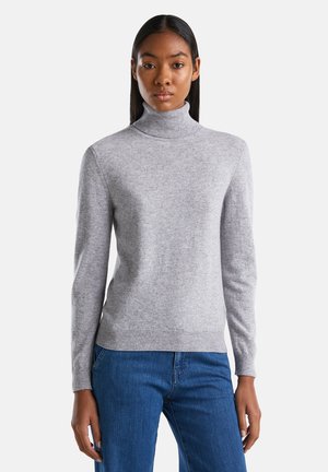 Femme portant un pull col montant gris clair et un jean bleu, se tenant de face devant un fond blanc uni.