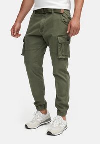 Homme portant un pantalon cargo vert olive avec plusieurs poches, des poignets élastiques, une ceinture assortie, et des baskets blanches à lacets.