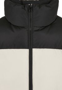 Padded Jacket mit hohem Kragen, Vorderreißverschluss und einem zweifarbigen Design in Schwarz und Creme. Der Stoff hat ein glattes, mattes Finish.