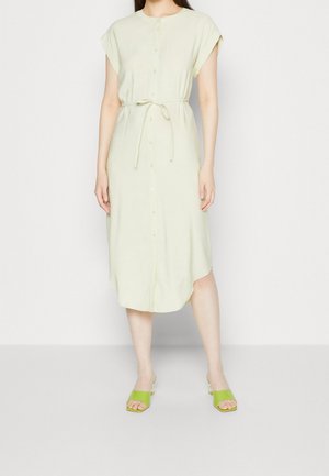 Kobieta ubrana w jasnofioletową, midi sukienkę zapinaną na guziki z podwiniętymi rękawkami i wiązaniem w talii, w połączeniu z limonkowozielonymi, odkrytymi sandałami na obcasie.