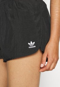 Ženská ruka spočívá vedle černých šortek Adidas s bílým logem u horní části stehna na bílém pozadí.