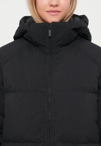 Veste matelassée noire avec un col haut, une fermeture éclair sur toute la longueur et un motif matelassé, avec une finition douce et mate et une capuche ajustée.