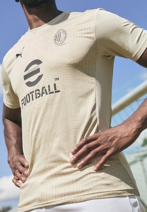 Persoon die een beige AC Milan voetbalshirt draagt met het Puma-logo en de sponsor "eFootball", staand in de buitenlucht met handen in de zij.