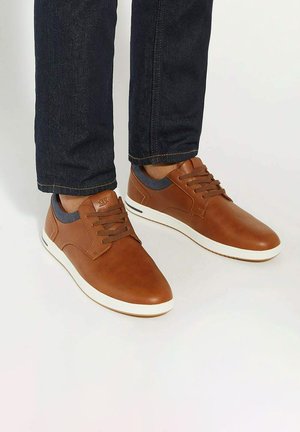 Bruine leren sneakers met grijze stofaccenten, witte rubberen zool en zes ogen voor veters, gecombineerd met een donker spijkerbroek.
