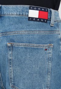 Artimas mėlyno džinsinio audinio kelnių užpakalinio kišenės vaizdas su Tommy Jeans etikete ir mažuoju raudonu, baltu bei tamsiai mėlynu logotipo pleistru kišenėje.