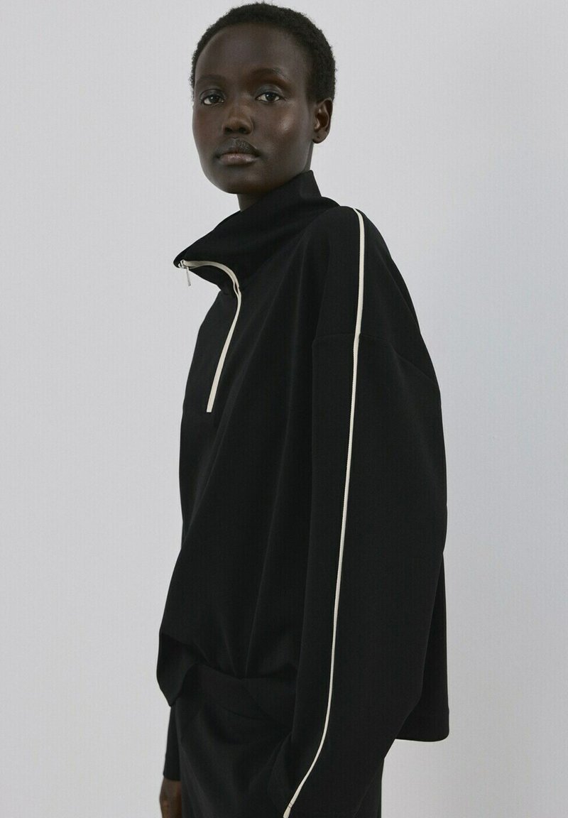 Jeune femme noire portant un pull-over noir ample avec une fermeture éclair blanche et des bordures blanches, debout devant un fond gris clair uni.