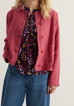 Femme portant une veste rouge boutonnée sur un chemisier foncé à fleurs avec un jean bleu, debout avec une main dans la poche sur un fond uni.
