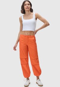 Orange cargo pants with outer pockets, drawstring closure, and tapered cuffs, paired with a white sleeveless top and white sneakers.