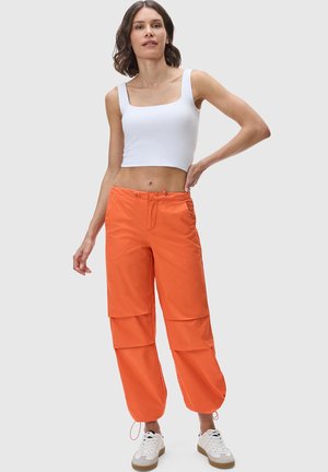 Orange Cargo-Hosen mit Außentaschen, Kordelzug-Verschluss und schmal zulaufenden Bündchen, kombiniert mit einem weißen ärmellosen Oberteil und weißen Sneakers.