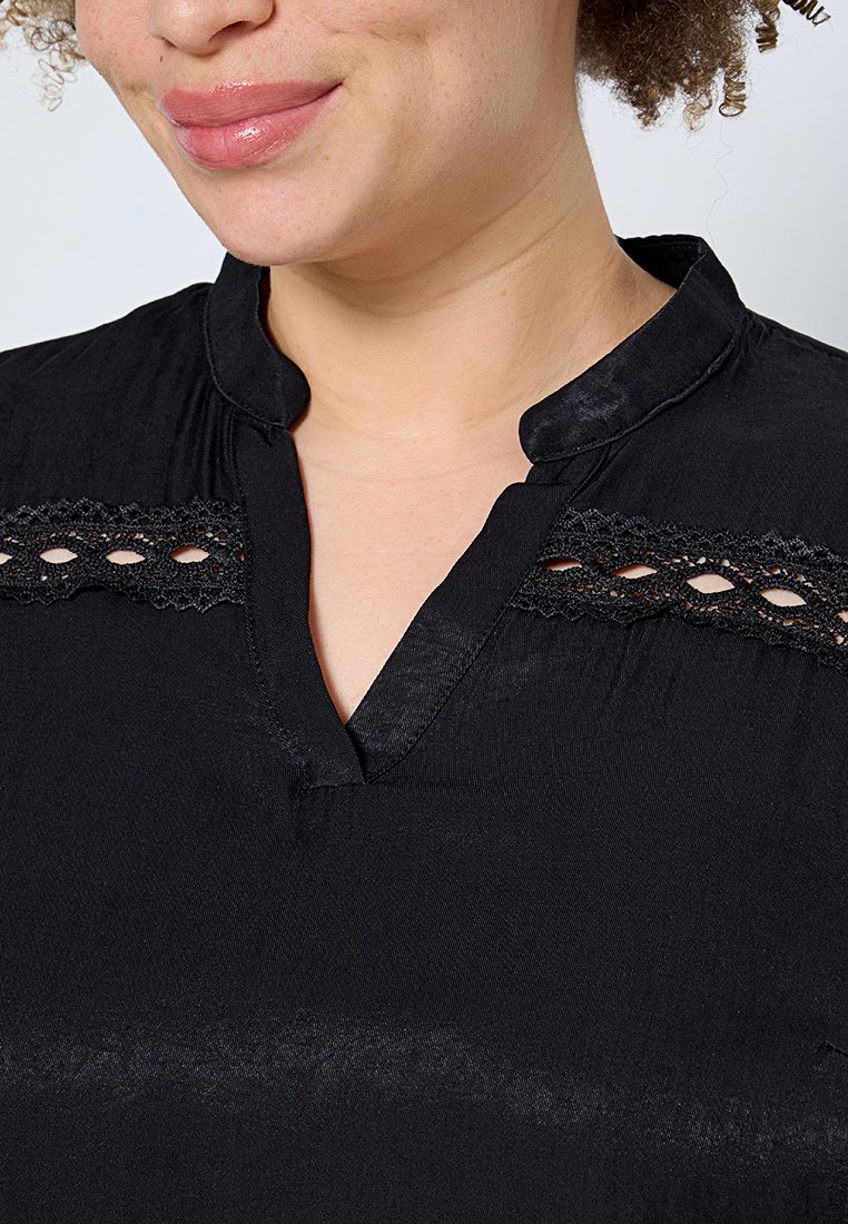 Blouse noire avec un léger lustre, décolleté en V et dentelle sur les épaules. Le tissu semble doux avec une texture lisse.