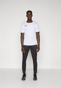Tricou sportiv alb cu mâneci scurte și logo-ul Puma negru, asortat cu pantaloni negri mulți și încălțăminte sport neagră.