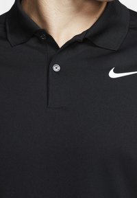 Detailní záběr na osobu, která má na sobě černé polo tričko s límečkem, knoflíkem a bílým logem Nike swoosh na pravé části hrudníku.