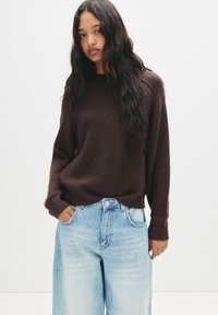 Maglione marrone a coste con maniche ampie, abbinato a jeans larghi a vita alta di colore blu chiaro. Il modello sta in piedi con le mani nelle tasche, rivolto in avanti.