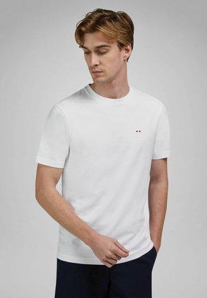 Joven con cabello castaño claro, vestido con una camiseta blanca de manga corta lisa y pantalones oscuros, mirando hacia abajo con una mano en el bolsillo.