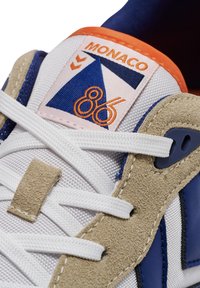 Sneaker met wit gaas en blauwe suède accenten, met een oranje en roze label waarop "MONACO 86" staat en een geometrisch ontwerp.