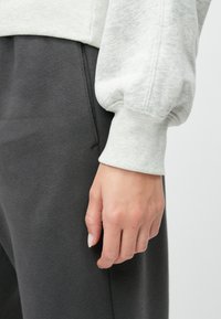 Svijetlosiva dukserica s rebrastim manžetama, uparena s tamnosivim trenirkama. Mekani materijal s glatkom teksturom. Ruka naslonjena na hlače.