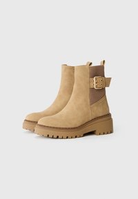 Bottines en suède beige avec un bout rond, un panneau élastique côtelé et une boucle sur le côté. Semelle en caoutchouc épaisse avec un profil texturé.