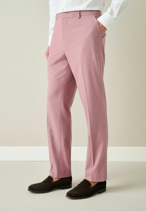 Mann trägt maßgeschneiderte staubrosa Hose, weißes Hemd und dunkelbraune Loafer, steht mit einer Hand in der Tasche vor schlichtem Hintergrund.