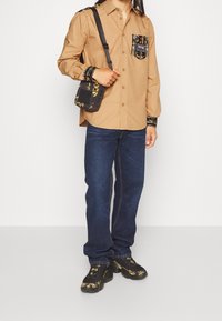 Brun skjorta med knappar och svarta mönstrade ärmar och ficka, i kombination med blå jeans och svarta skor med gula blommiga accenter. Svart axelväska.