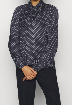 Blouse marine avec un col à nœud, présentant un motif géométrique blanc, des manches longues et des poignets à boutons, fabriquée en tissu léger.