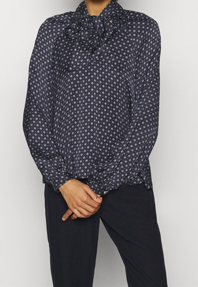 Blusa blu navy con colletto a fiocco, caratterizzata da un motivo geometrico bianco, maniche lunghe e polsini con bottoni, realizzata in tessuto leggero.