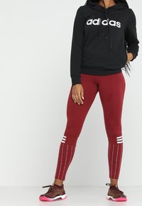 Felpa nera con logo "adidas" bianco, leggings bordeaux con strisce bianche e dettagli riflettenti, sneakers marroni con accenti rosa.