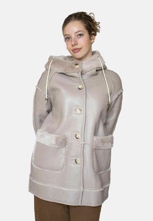 Nuori nainen, jolla on päällään beige hupullinen lammasturkistakki, jossa on suuret taskut ja napit, seisoo yksinkertaisen valkoisen taustan edessä.