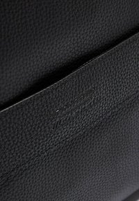 Sac en cuir noir à texture grainée, présentant un design épuré et un logo de marque embossé sur le devant. Les coutures sont discrètes et raffinées.