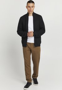 Sort zip-up trøje med teksturerede ærmer, hvid T-shirt, brune bukser og sorte sneakers med hvide såler; modellen står i en neutral indstilling.