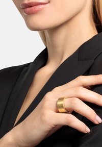 Anello color oro con una fascia liscia e ampia, con dettagli incisi, indossato su una mano con manicure naturale, su una giacca nera.