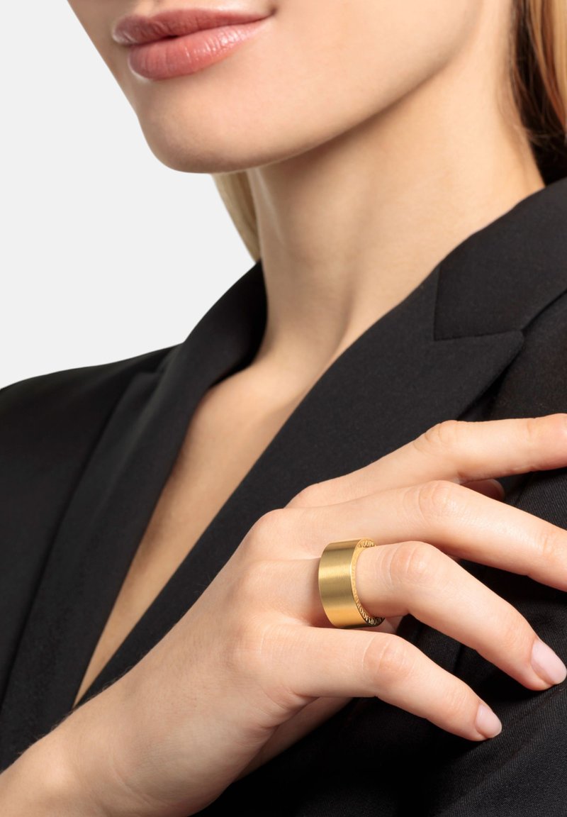 Anello color oro con una fascia liscia e ampia, con dettagli incisi, indossato su una mano con manicure naturale, su una giacca nera.