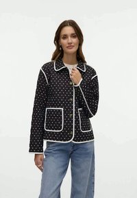 Casaco preto acolchoado com bolinhas brancas, com acabamento em branco em contraste, bolsos frontais e botões dourados. Usado sobre uma blusa de cor clara.
