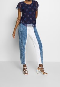 Navy floral-patterned top with sheer sleeves paired with color-blocked denim jeans, featuring light blue and white segments, and white strappy heels.