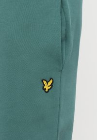 Παντελόνια φόρμας σε τιρκουάζ χρώμα από μαλακό ύφασμα, με κίτρινο έμβλημα πουλιού με μαύρο περίγραμμα στην αριστερή πλευρά κοντά στην τσέπη.