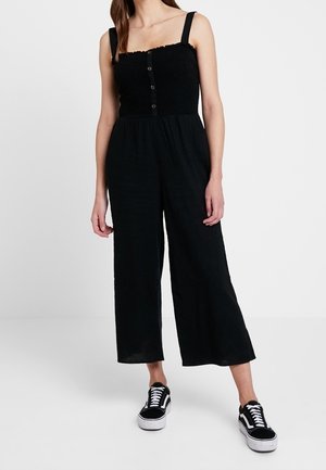 Mono negro con corpiño fruncido, escote cuadrado y tirantes ajustables. Cuenta con botones frontales y silueta de pierna ancha. Combinado con zapatillas negras.