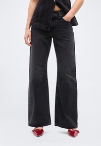 Άτομο που φοράει ψηλόμεσα μαύρα τζιν με φαρδιά μπατζάκια, μαύρο αμάνικο τοπ και κόκκινα παπούτσια μυτερά, στέκεται μπροστά σε απλό φόντο.