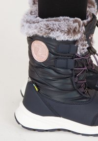 Sorte vinterstøvler med en fyldig grå fake fur kant, udstyret med et rundt pink logo-patch og lilla snørebånd, placeret på en hvid gummisål.