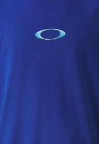 Zila kokvilnas T-kreklus ar gludu tekstūru. Centrā ir viegli zils ovāls logotips izšūts. Vienkāršs, apaļais kakla dizains.